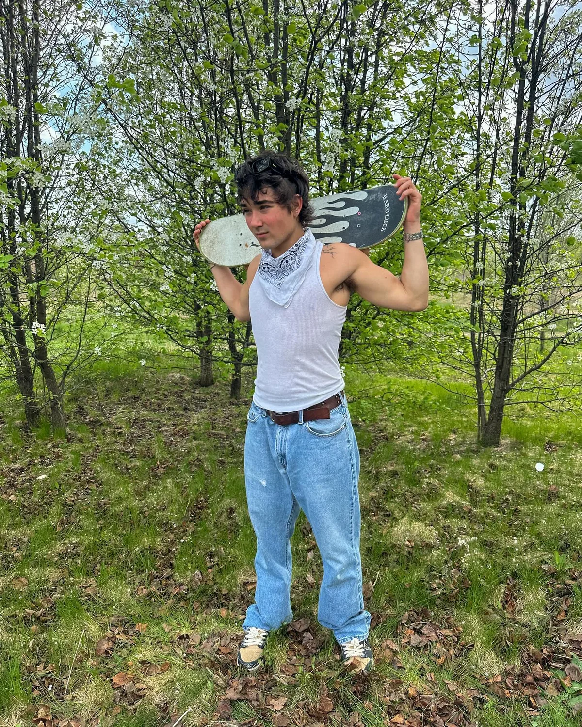 Alex carrying a skateboard across his shoulders among green trees