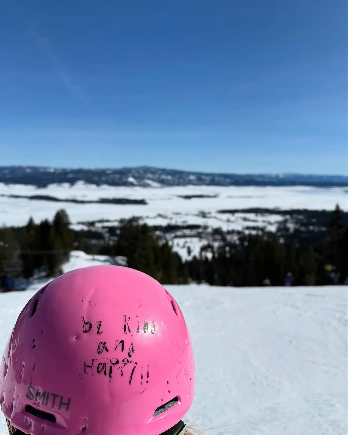 A pink helmet with a handwritten Be Kind and Happy message above a snowy mountain view